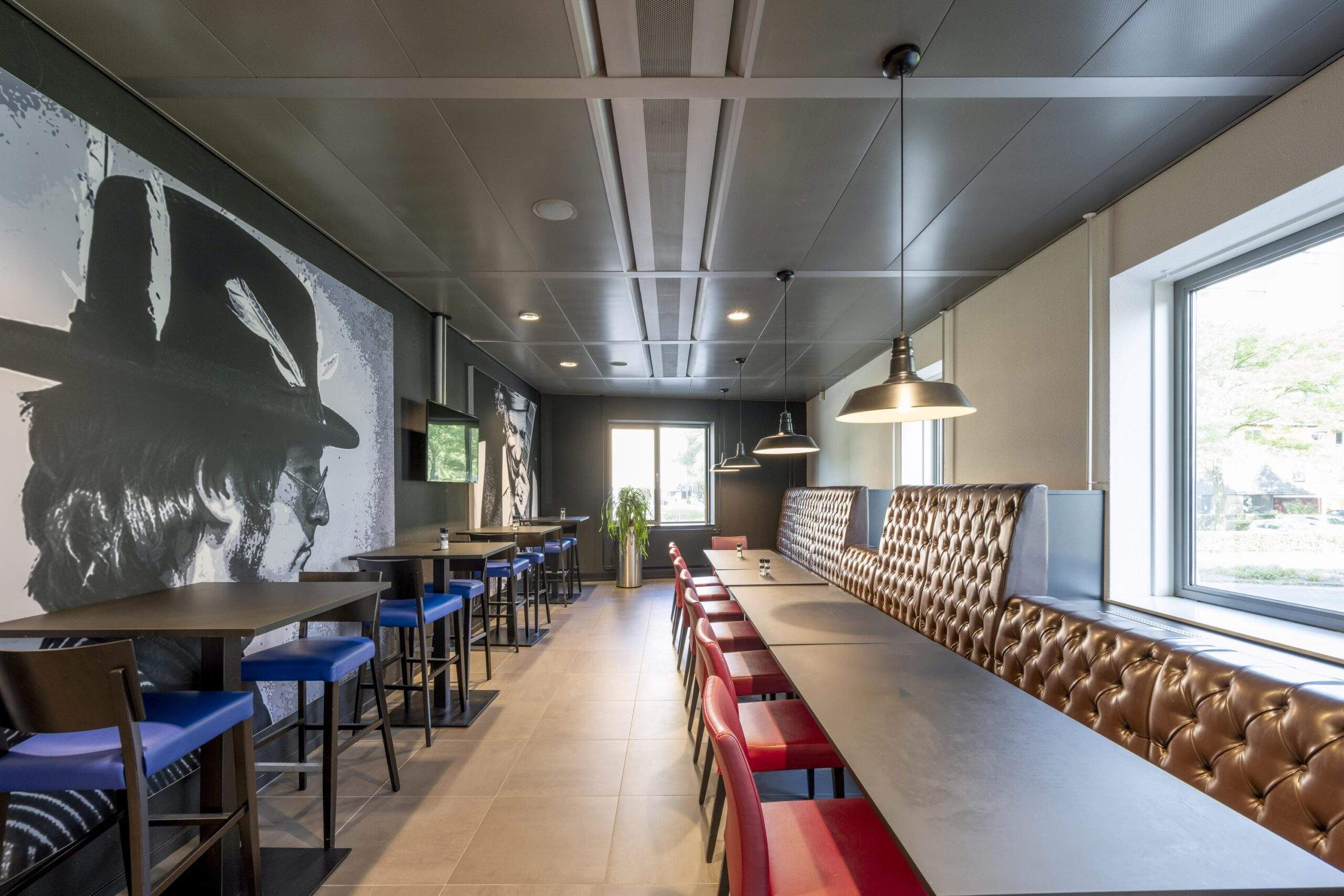 Modern ingerichte eet- of werkruimte aan het Koningin Wilhelminaplein met lange tafels, gekleurde stoelen, kunst aan de muur en grote ramen.